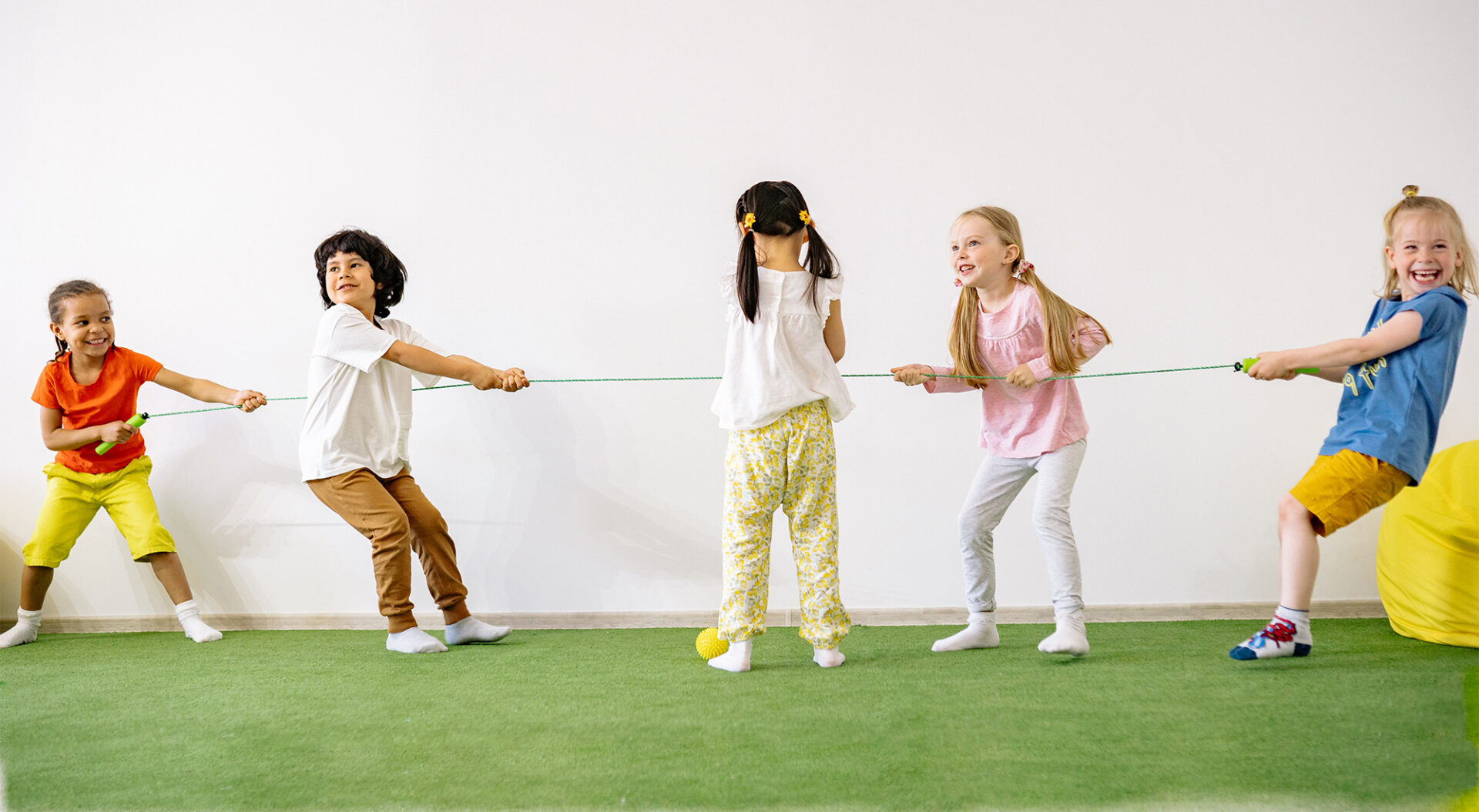 Vier lachende Kinder spielen mit einem Springseil Tauziehen, ein Kind steht in der Mitte