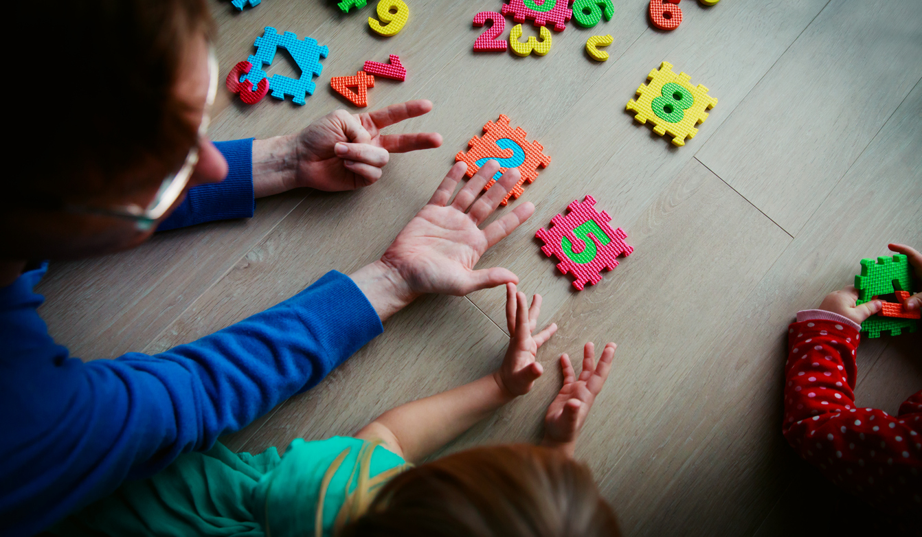 Eine erwachsene Person und zwei Kinder spielen mit einem Ziffernpuzzle und zeigen Ziffern mit den Fingern