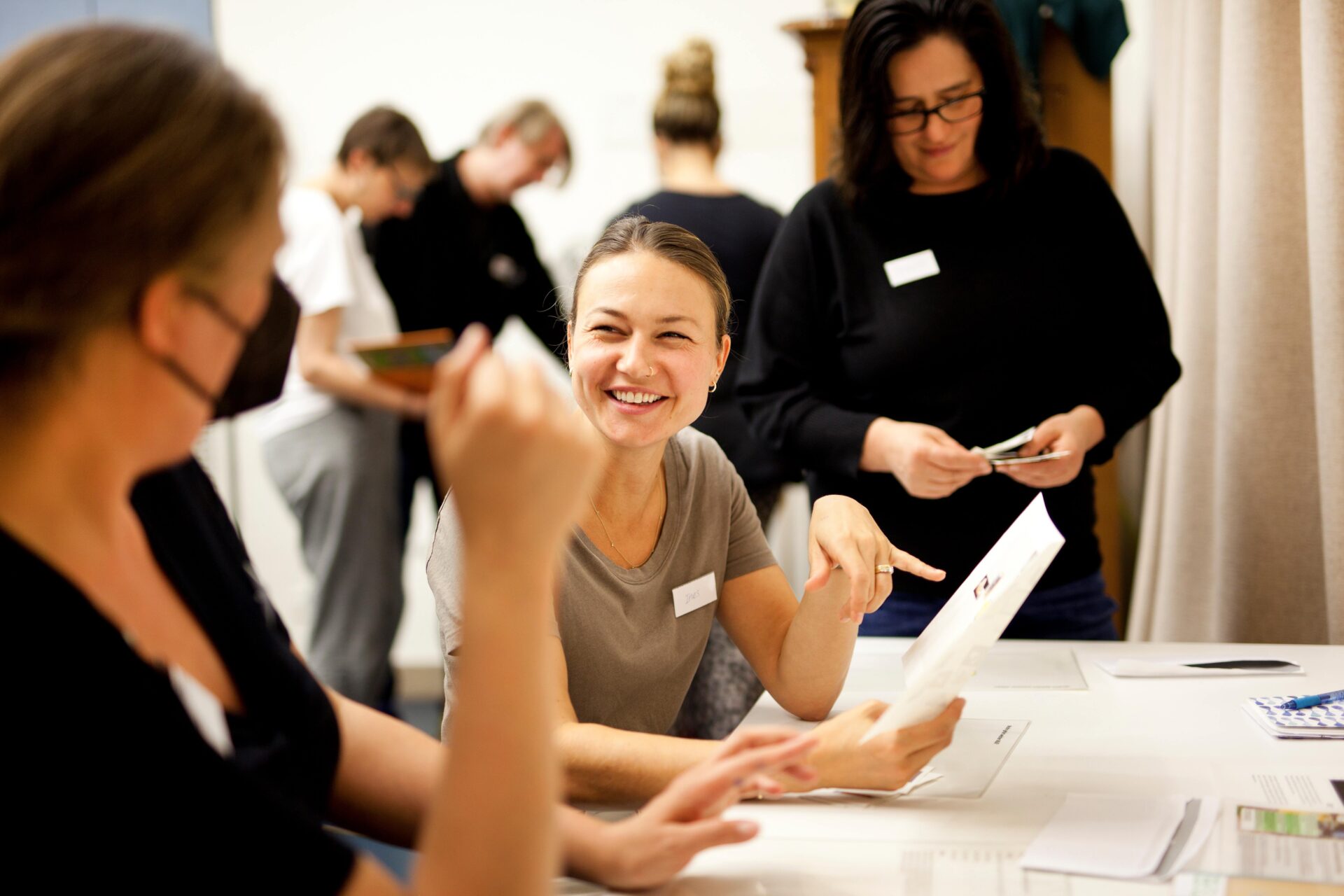 Zwei Personen befinden sich in einer Unterhaltung. Eine Person ist von hinten zu sehen, eine Person ist von vorne zu sehen, sie lacht und zeigt auf ein Blatt Papier, was sie in der Hand hält. Im Hintergrund blickt eine weitere Person auf Karten, die sie in der Hand hält.